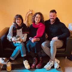 Lea, Shadia und Daniel sitzen auf einer schwarzen Ledercouch und lachen in die Kamera. Auf der Lehne hinter ihnen steht das Maskottchen des Kinderbüros: eine Kuscheltiergiraffe. Lea hält einen Kuscheltierwolf in den Händen.